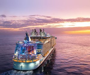 OA, Oasis of the Seas, aerial shot of ship at sea, sunset, dusk, evening, 3/4 aft starboard view, ocean horizon, red, orange, purple colors, AquaTheater and slides at rear of ship, scenic, seascape